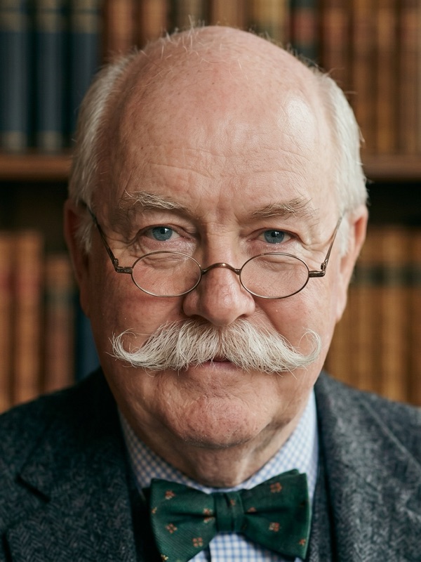 Professor Cornelius Underwood, an older distinguished man with a white walrus mustache, wire-rim half-moon spectacles, a charcoal tweed suit and a green bow tie, in a warm library setting.